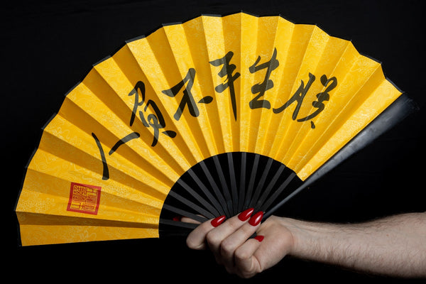 Bright yellow folding hand fan with black bamboo ribs and calligraphy, held open against a black background